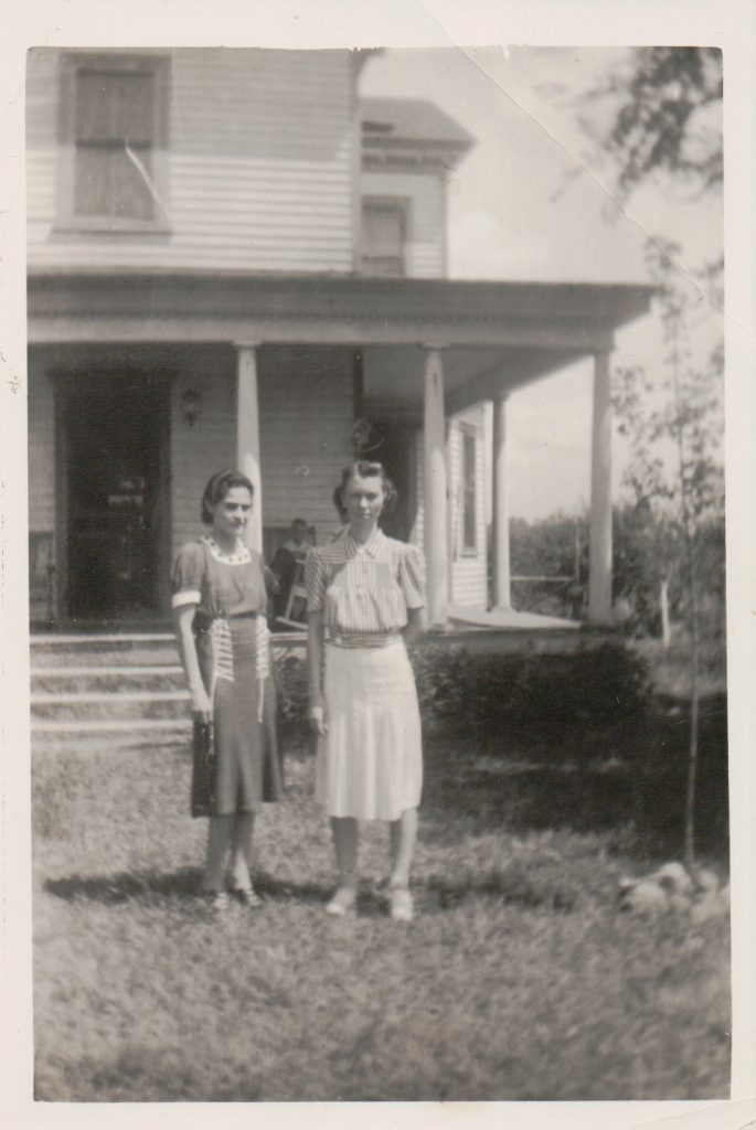 Black and white photo of 2 young women standing in front of a large 2 story house.