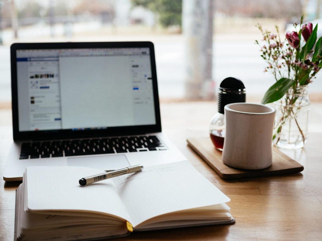 laptop and open notebook with pen on a table with a coffee mug beside it.